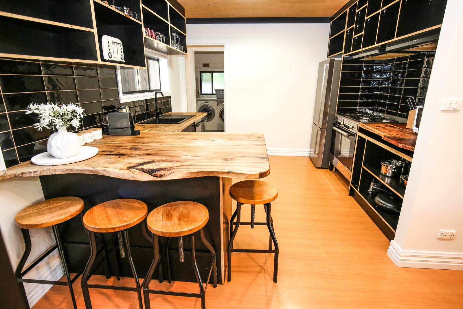 The kitchen: a timber-slab island bench with stools, black tiled walls and open timber shelving