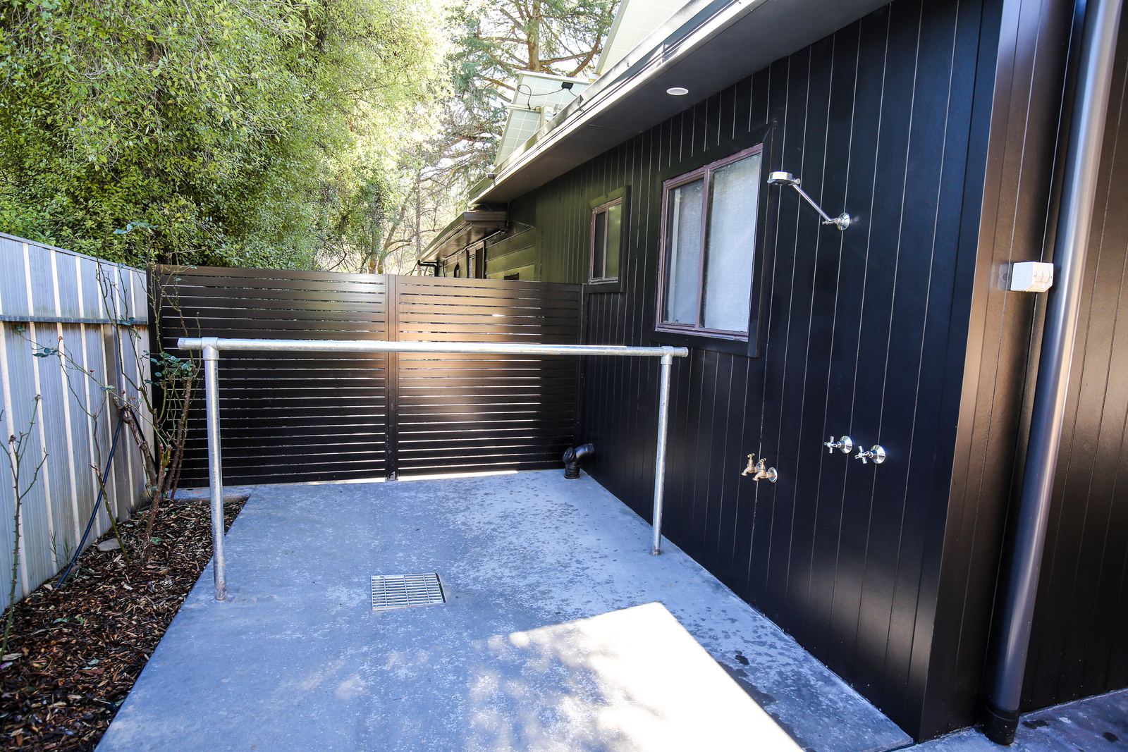 The outdoor shower on the black-clad wall beside the house, with the drying rail in front of it